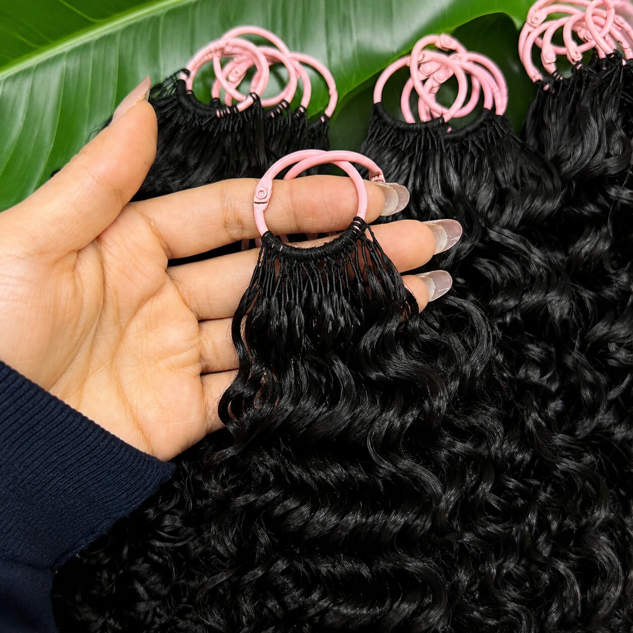 Close-up of a Black woman holding a single bundle of Burmese curly crochet hair, showing natural curls and shine, pink loop attachment visible, green leaf background.
