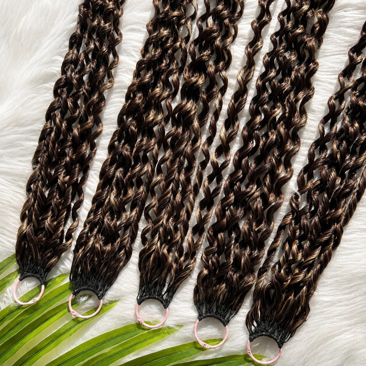 Top-down display of multiple Feathered Crochet Hair bundles with deep brown and caramel highlights, coiled feathered curls, shiny and smooth texture on white faux-fur background with tropical leaves