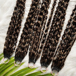 Top-down display of multiple Feathered Crochet Hair bundles with deep brown and caramel highlights, coiled feathered curls, shiny and smooth texture on white faux-fur background with tropical leaves