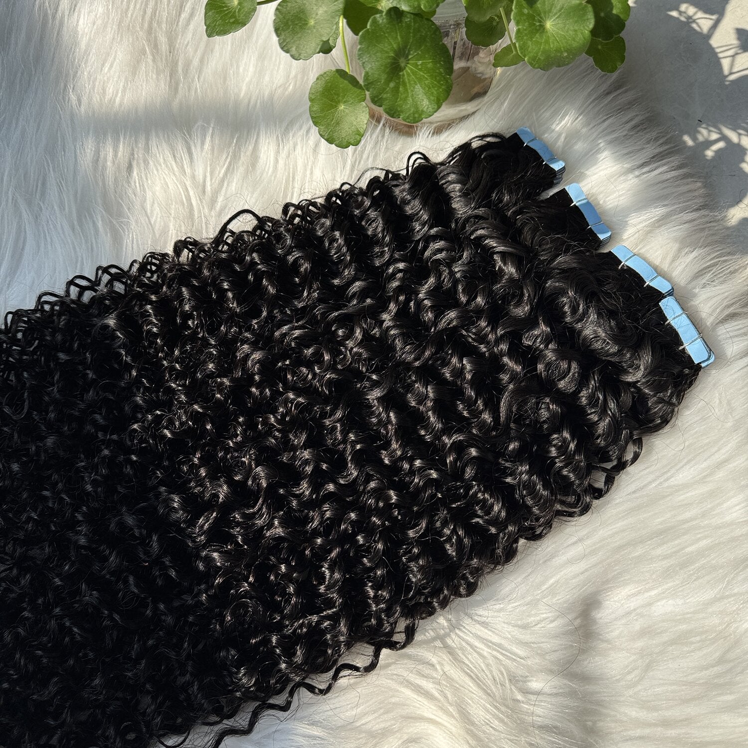 Top-down flat lay of curly black tape-in hair extensions arranged neatly on a soft white faux-fur background. Icons highlight product features: 100% virgin human hair, lightweight & comfortable, reusable, heat styling friendly, strong tape for long-lasting hold.